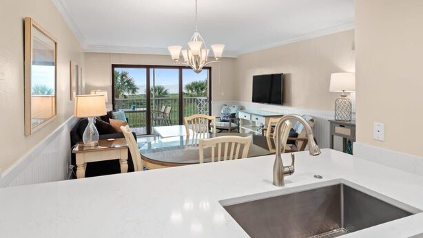 A modern kitchen with a central island, a dining area with chairs, a TV mounted on the wall, and a balcony with outdoor furniture.