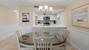 A dining area with a round glass table, four chairs, a chandelier, a kitchen in the background, and framed artwork on the walls.