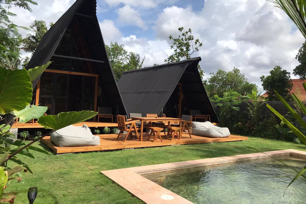 A modern A-frame structure with a pool, wooden deck, and outdoor seating area.