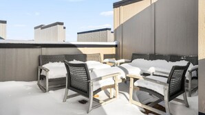 A rooftop patio with wicker chairs and tables covered in snow, against a backdrop of a clear sky and building rooftops. - Downtown TC Condo | Rooftop + EV Charger Cornhole (Traverse City)