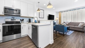 A modern kitchen with white cabinets, stainless steel appliances, and a central island. There is a dining area with a blue chair and a sofa. - Downtown TC Condo | Rooftop + EV Charger Cornhole (Traverse City)