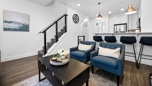 A modern living room with a dark wood floor, a black coffee table, and blue armchairs. A staircase with black railings leads to a kitchen area with white cabinets and a stainless steel refrigerator. - Downtown TC Condo | Rooftop + EV Charger Cornhole (Traverse City)
