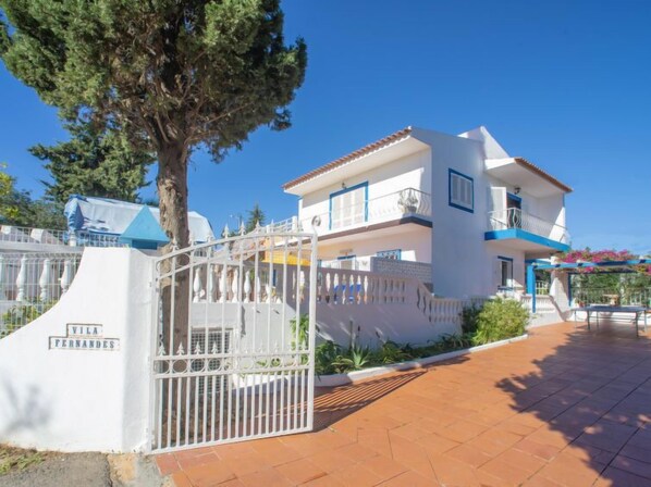 A two-story villa with a white exterior and blue accents, a gated entrance, and a paved driveway.