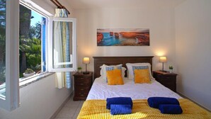 A bedroom with a bed, two bedside tables, a window with curtains, a painting on the wall, and a view of greenery outside.