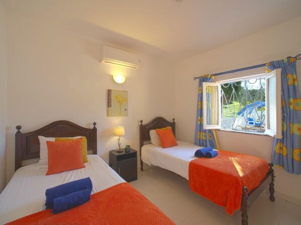 A room with two beds, each with a wooden headboard, white bedding, and orange and blue towels. There is a nightstand with a lamp and a small table with a plant. A window with blue curtains and a view of a blue car outside is visible.