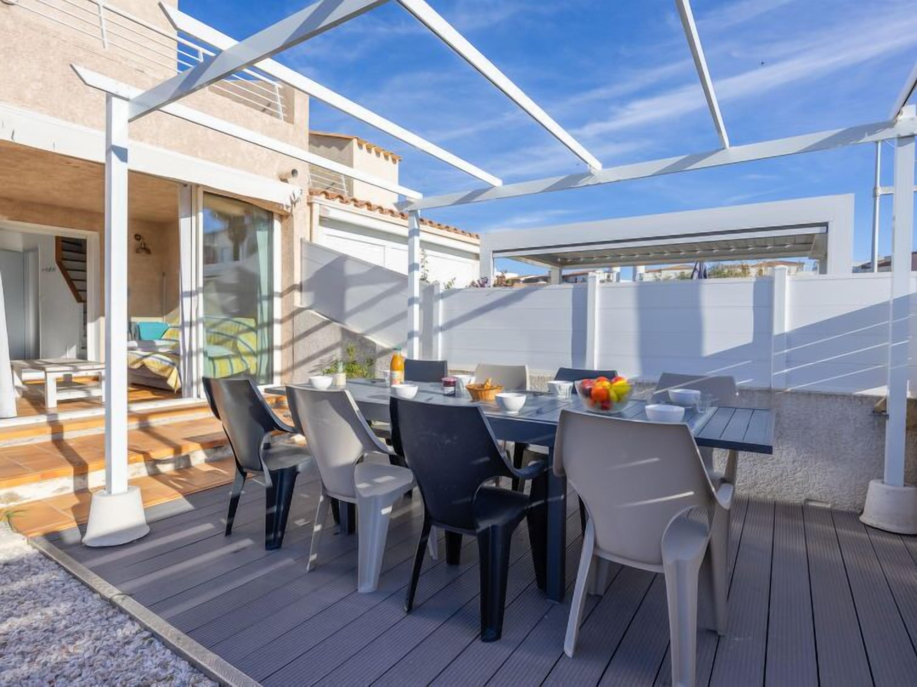 An outdoor patio with a table set for dining, surrounded by chairs, and a covered area providing shade.