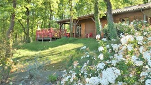 A rustic cabin with a wooden deck and a covered porch, surrounded by lush greenery and white flowers. - Le Chêne Vert by Interhome (Loupiac)