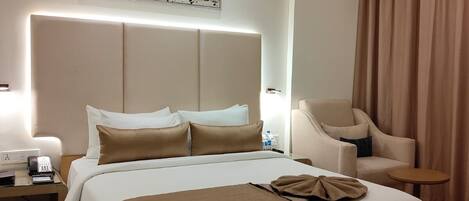 A modern hotel room with a large bed, a bedside table with a phone, a chair, a small table, and a painting on the wall.