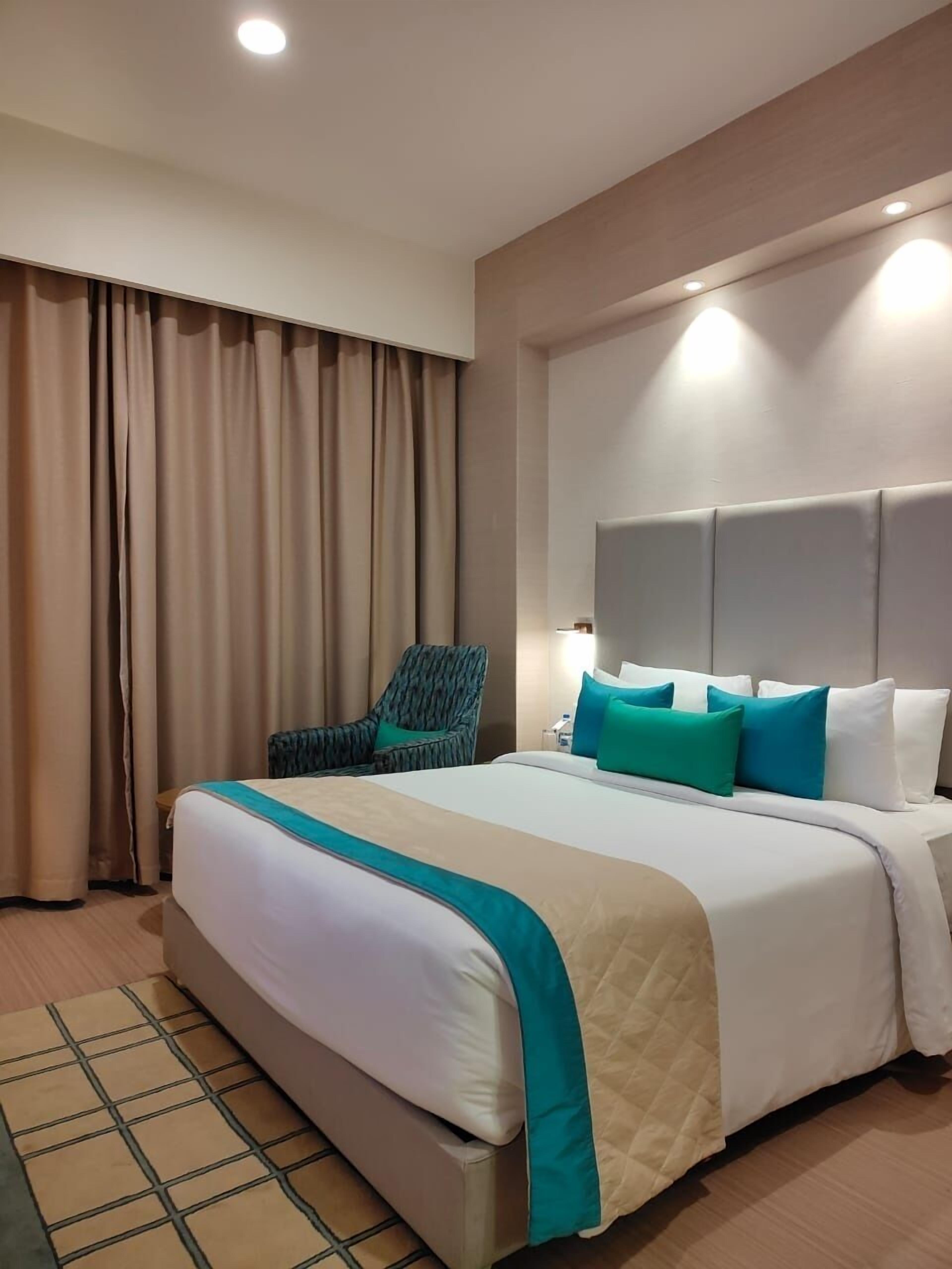 A hotel room with a large bed, a chair, a nightstand, and a wall-mounted lamp.