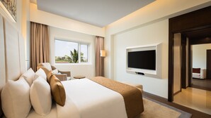 A hotel room with a large bed, a flat-screen TV, a window with curtains, and a view of palm trees. - Fortune Hosur  -  Member ITC Hotels' Group (Hosur)