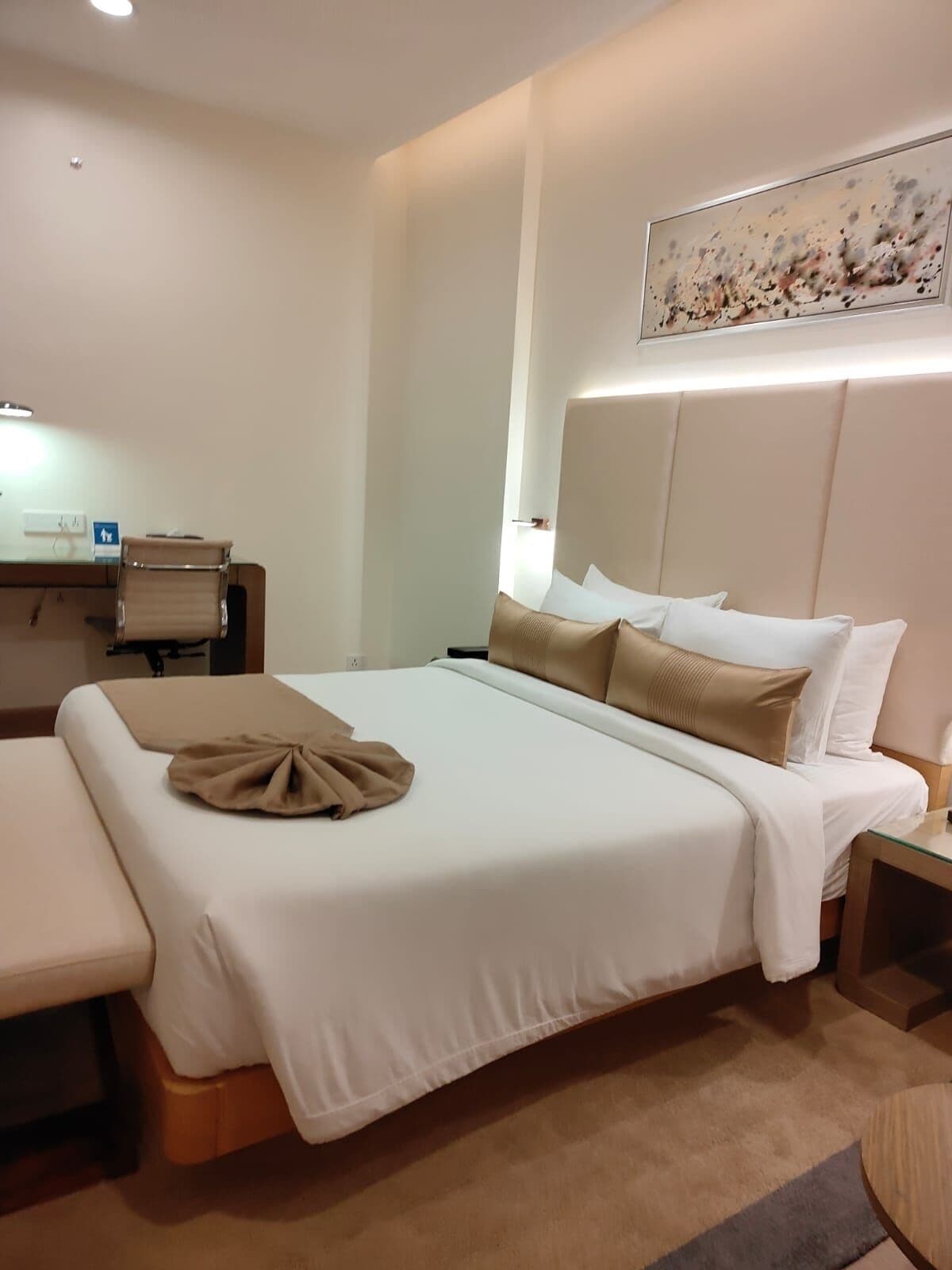 A hotel room with a large bed, a bedside table, a desk with a chair, and a painting on the wall.