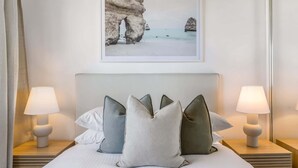Una cama con cabecera, almohadas, lámparas de noche y un cuadro con una escena costera.