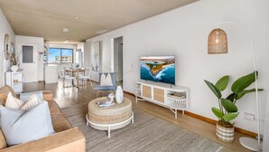 Amplia sala de estar con televisión, reposapiés de mimbre y comedor con mobiliario blanco.