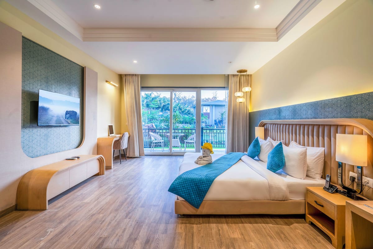 A modern hotel room with a large bed, a desk, a seating area, and a balcony with a view of greenery.