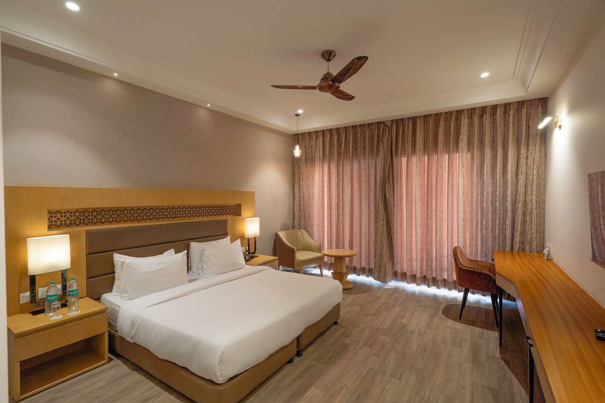 A hotel room with a large bed, a desk, a chair, a nightstand with a lamp, and a ceiling fan.