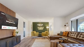 A modern living room with a large flat-screen TV, a sofa, a coffee table, and a green accent wall with a neon sign. - Evergreen Luxe! Backyard Oasis w/ Pool & Hot Tub! (Colorado Springs)