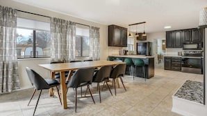 A modern kitchen with dark cabinets, a wooden dining table, and a large window with curtains. - Evergreen Luxe! Backyard Oasis w/ Pool & Hot Tub! (Colorado Springs)