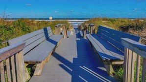 Une promenade en bois menant à une plage, avec des bancs et un panneau indicateur.
