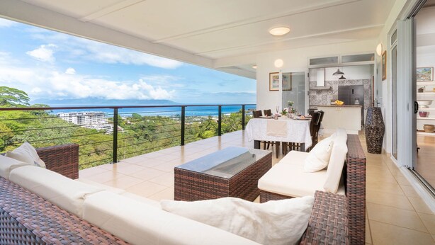 A balcony with wicker furniture, a dining table, and a kitchen area with a view of the ocean.
