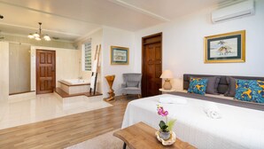 A hotel room with a bed, a wooden side table with a plant, a grey armchair, a small round table with a lamp, and a painting on the wall.