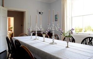 Comedor con una mesa larga dispuesta para seis personas, rodeada de sillas. Hay seis velas altas sobre la mesa, y un jarrón con flores moradas en el lado derecho.