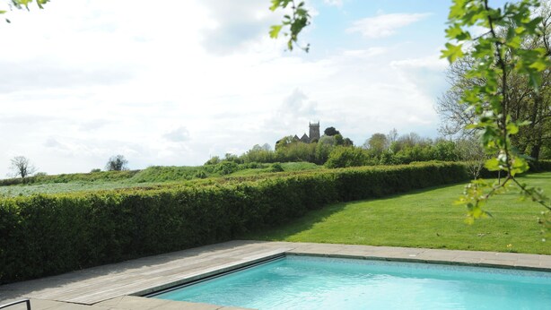 A swimming pool with a wooden deck, surrounded by a well-manicured lawn and a hedge. In the background, there is a hill with a building on top. - The Old Rectory with Swimming Pool (Bristol)