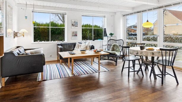 A spacious living room with a wooden floor, a black sofa, a dining table with chairs, and large windows offering a view of the outdoors.