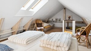 A loft bedroom with a sloped ceiling, two skylights, a bed with a white floral duvet, a wicker chair, and a small table with a picture frame.