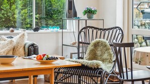 A cozy room with a large window, a wicker chair, a small table with a bowl of fruit, and a potted plant on a shelf.