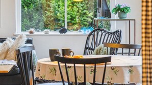 A dining area with a table set for two, chairs, a window with a view of greenery, and a hanging lamp.