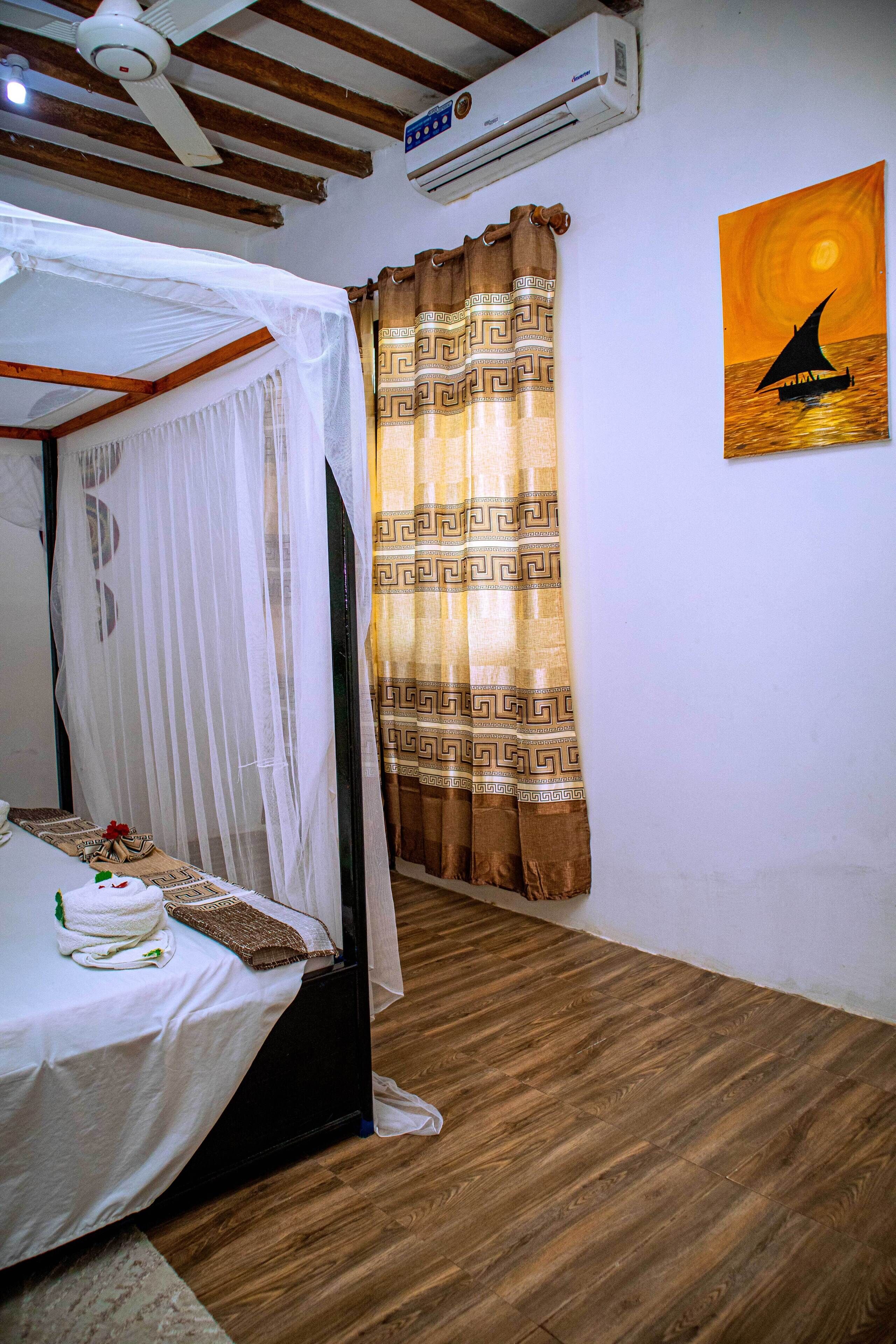 Une chambre à coucher avec un lit, un ventilateur de plafond, un climatiseur, des rideaux à motifs et un tableau représentant un coucher de soleil accroché au mur.