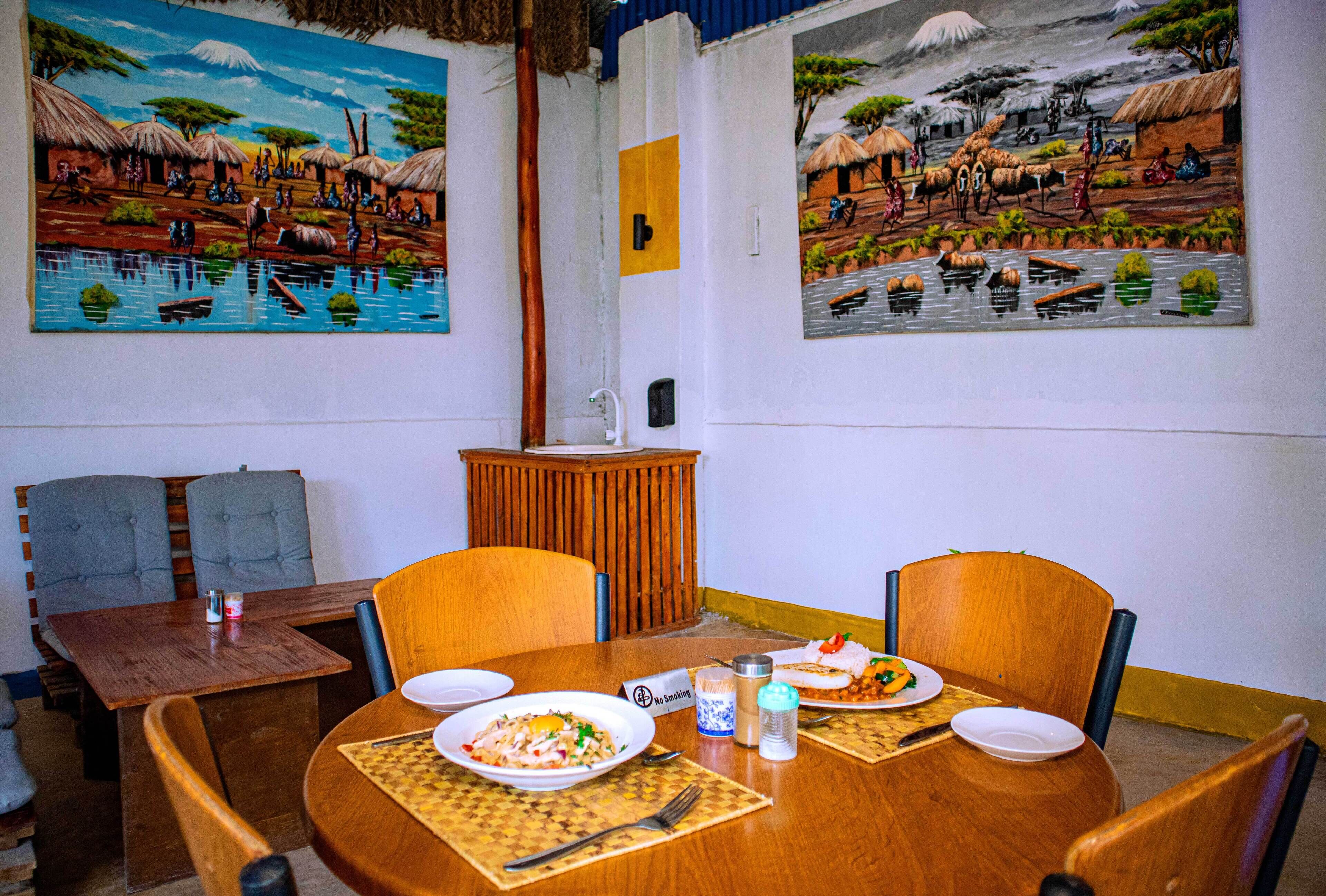 A dining area with a wooden table set for two, a small wooden cabinet, and two paintings on the wall depicting rural scenes.
