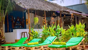 Un espacio lounge al aire libre con ambiente tropical, con tumbonas de madera, techo de paja y abundante vegetación.