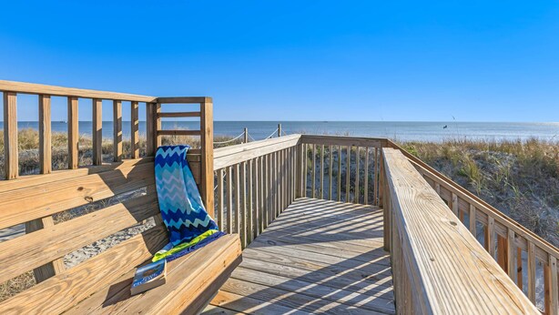 A wooden deck with a bench and a blanket, overlooking a beach with dunes and the ocean.