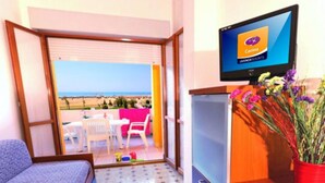 Ein Zimmer mit Balkon, einem Fernseher, der 'Carina' zeigt, einem kleinen Tisch mit Stühlen und einer Holzkommode mit Blumen.