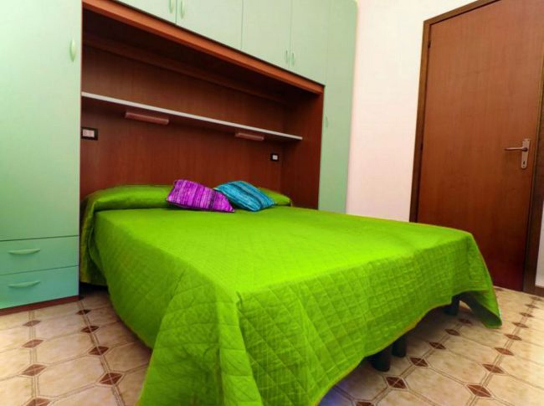 A small bedroom with a bed, a green bedspread, a wooden wardrobe, and a door.