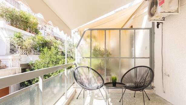A balcony with two chairs and a small table, a glass partition, and a view of greenery and buildings.