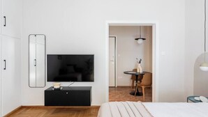 A modern hotel room with a large flat-screen TV mounted on the wall, a black console table, a bed with a white comforter, and a small dining area with a table and chairs.