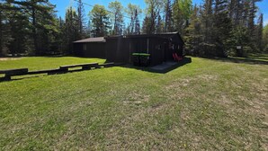 Une cabane en bois rustique, dotée d’un petit porche et d’une table de pique-nique, se trouve dans un espace verdoyant, entouré d’arbres.