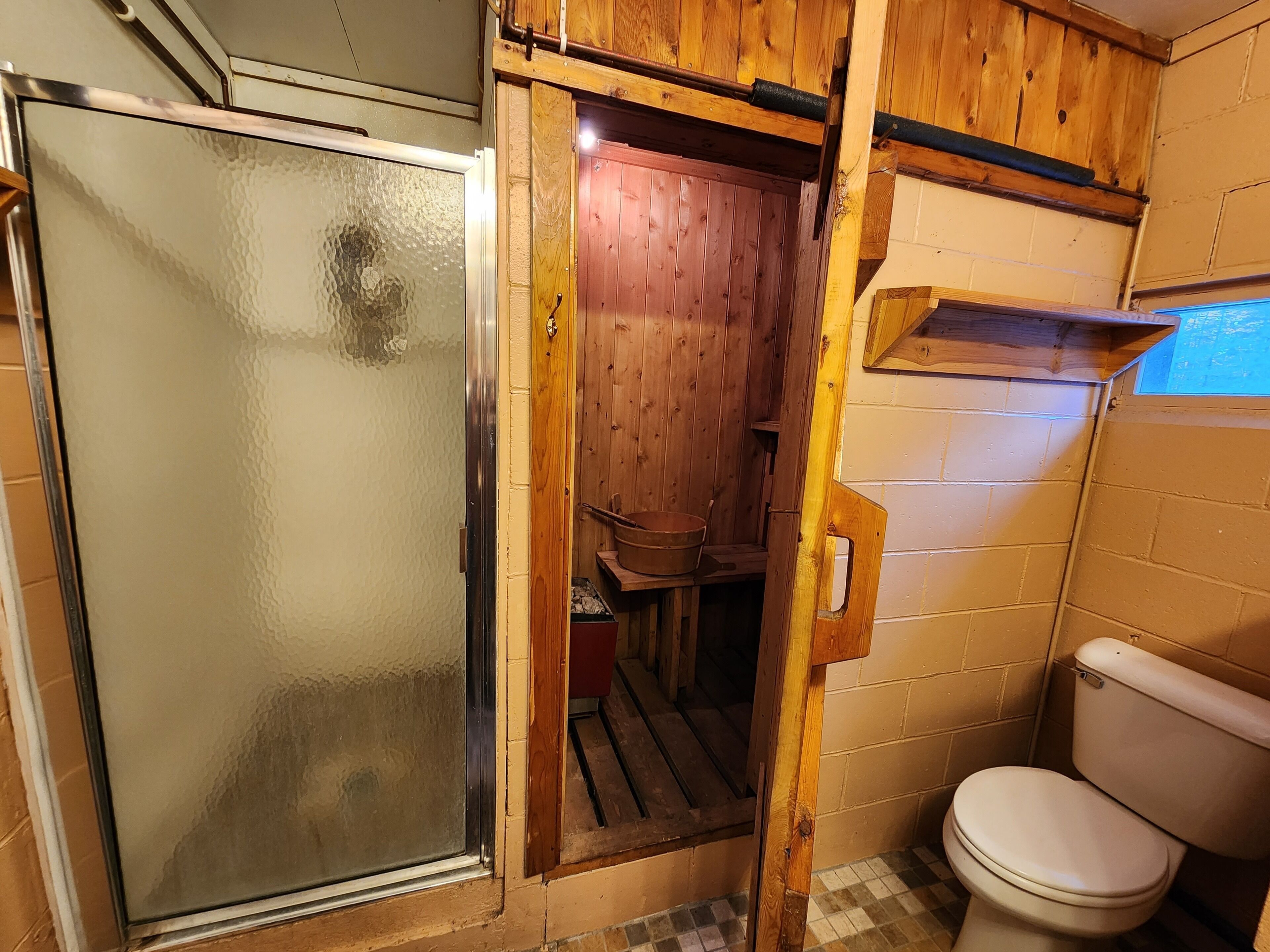 Et lille badeværelse med glasbrugedør, trævægge, toilet og en hylde.