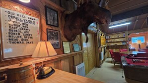 Un intérieur rustique avec une tête de wapiti accrochée au mur, des murs en bois, un coin bar et un tableau affichant une liste de noms.