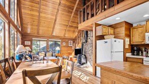 A wooden cabin interior with a dining area, kitchen, and a stone fireplace. - Fantastic Coastal Retreat in Wooded Setting, Pet Friendly, Short Walk to Beach, Huge Deck and Games (Cannon Beach)