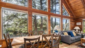 A spacious wooden cabin with large windows offering views of a forest. Inside, there is a dining table with chairs, a gray sofa, and two lamps. - Fantastic Coastal Retreat in Wooded Setting, Pet Friendly, Short Walk to Beach, Huge Deck and Games (Cannon Beach)