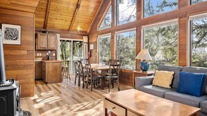 A cozy cabin interior with a dining area, kitchen, and a sofa by a large window. - Fantastic Coastal Retreat in Wooded Setting, Pet Friendly, Short Walk to Beach, Huge Deck and Games (Cannon Beach)