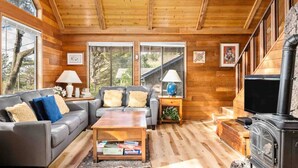 A cozy log cabin interior with a fireplace, wooden floors, and large windows offering a view of the outdoors. - Fantastic Coastal Retreat in Wooded Setting, Pet Friendly, Short Walk to Beach, Huge Deck and Games (Cannon Beach)