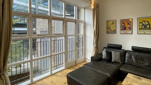 A modern living room with a large window, a black leather sofa, a wooden coffee table, and a hanging light fixture. - Stunning 3BD Maisonette W/balcony - Old Street! (London)