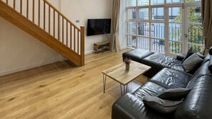 A spacious living room with a wooden staircase, a leather sofa, a coffee table, and a large window with curtains. - Stunning 3BD Maisonette W/balcony - Old Street! (London)