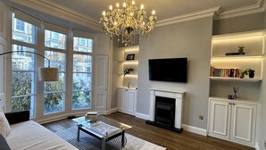 A living room with a fireplace, a chandelier, a flat-screen TV, and bookshelves. - Elegant & Beautifully Presented 1BD Flat- Chelsea! (London)