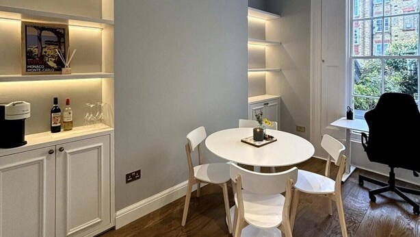 A modern dining area with a round table, white chairs, and built-in shelves with lighting. - Elegant & Beautifully Presented 1BD Flat- Chelsea! (London)