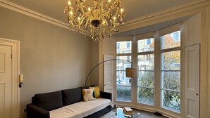 A living room with a dark sofa, a glass coffee table, and a large window with a view of buildings and trees. - Elegant & Beautifully Presented 1BD Flat- Chelsea! (London)